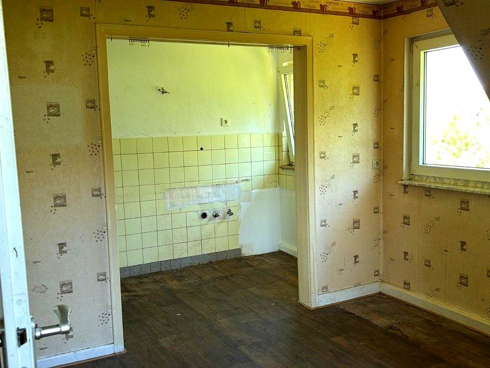 Wohnraum vor der Sanierung mit alter Tapete und Küchendurchgang